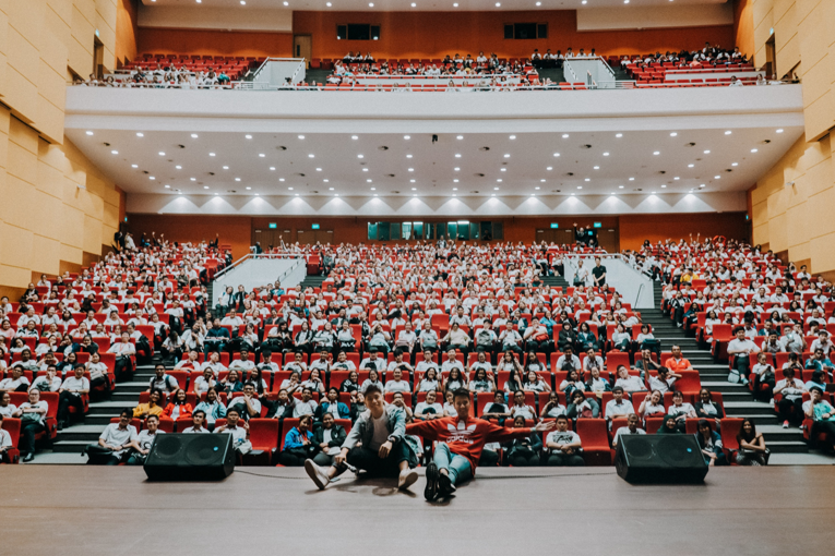 A large auditorium filled with seated people; three individuals are on stage near two large speakers.